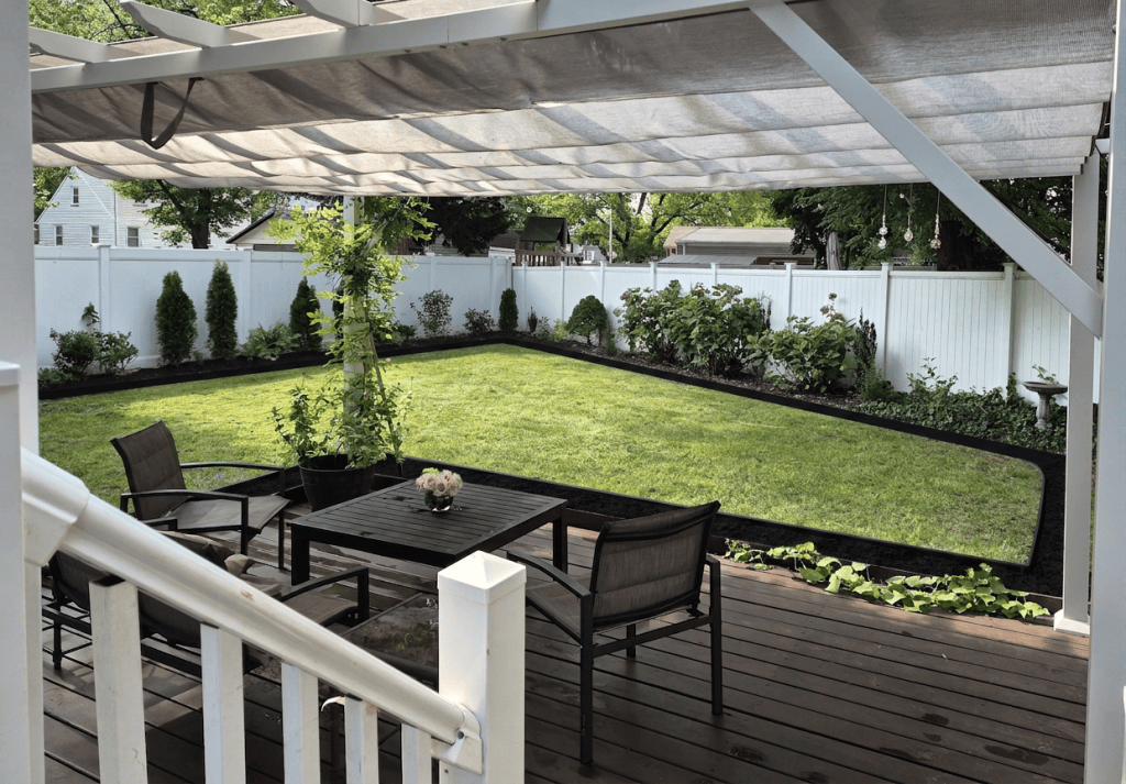 Backyard garden viewed from a covered deck, with patio furniture, a lawn, white privacy fence, flowering shrubs, evergreens, and garden beds.
