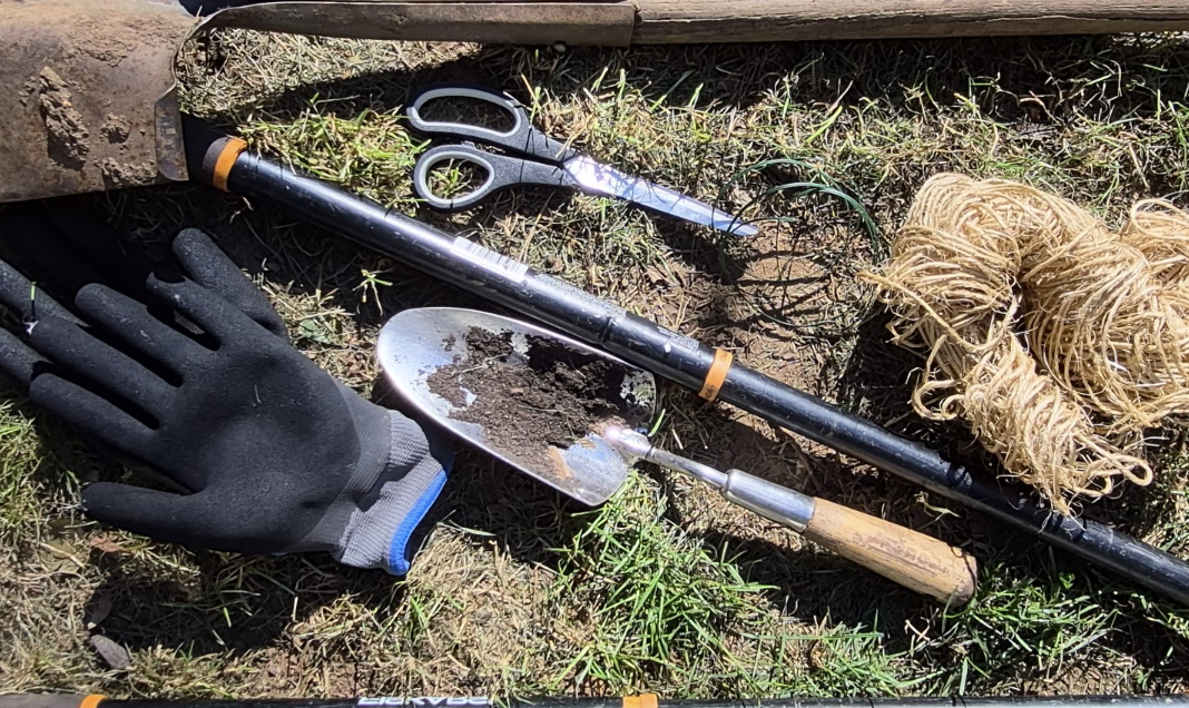 Garden maintenance tools including gloves, pruning shears, a trowel, twine, and long-handled tools laid out on the grass.