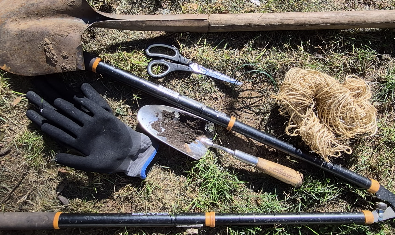 Garden maintenance tools including gloves, pruning shears, a trowel, twine, and long-handled tools laid out on the grass.