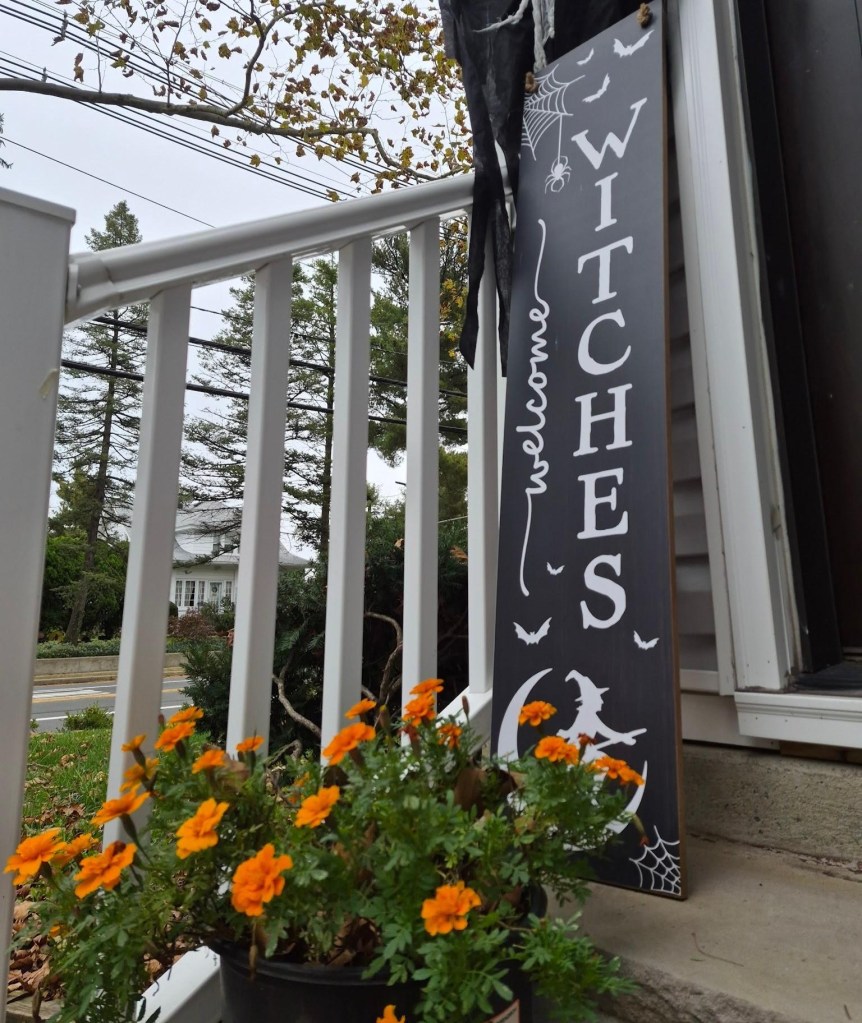 Orange marigolds bloom on a front porch beside a black Halloween decoration featuring the word WITCHES.
